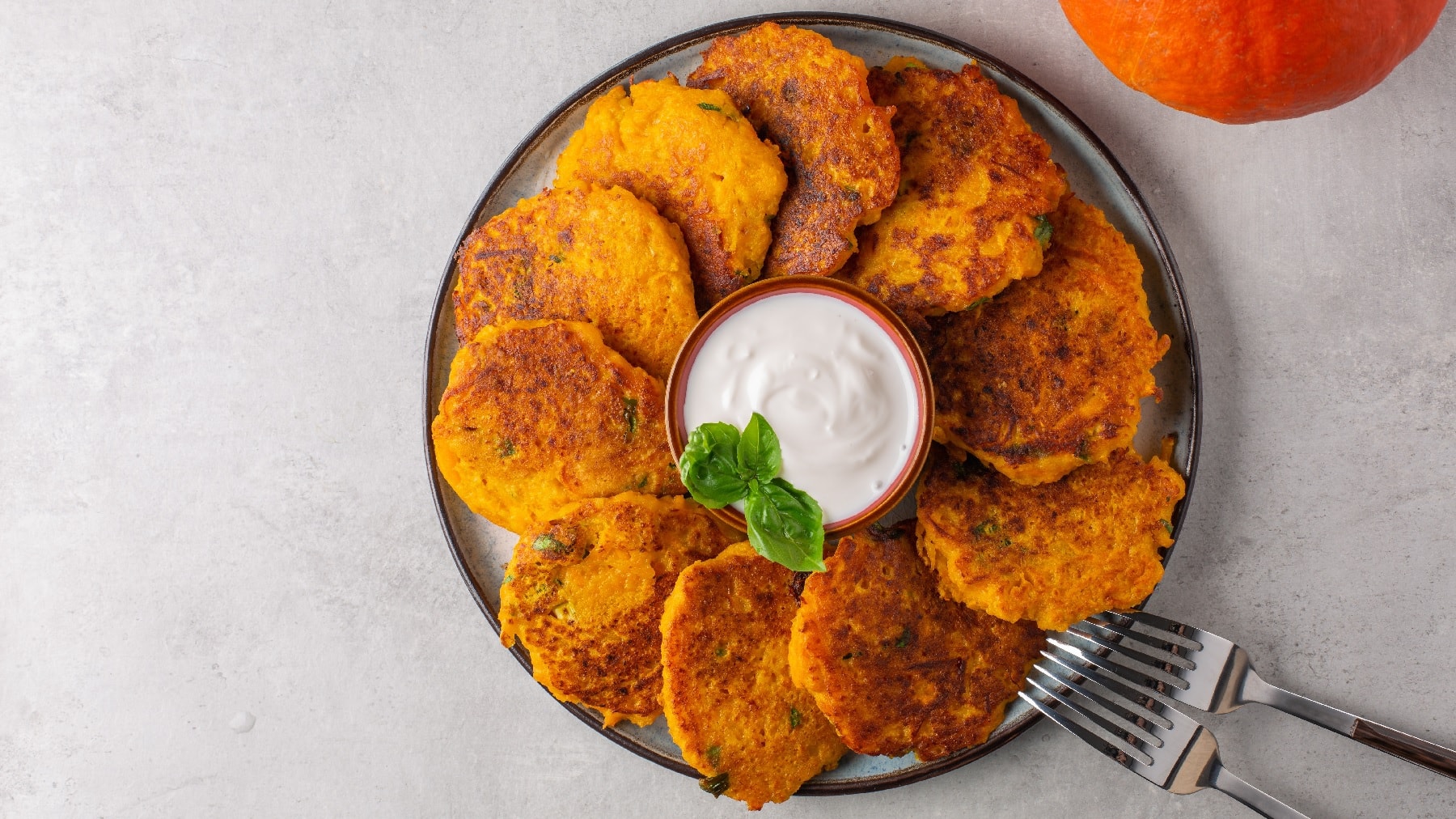 Mehrere Kürbis-Hashbrowns auf einem Teller übereinanderliegend im Kreis angeordnet und in der Mitte eine kleine Schale mit einem weißen Dip. Alles auf einem hellgrauen Untergrund.