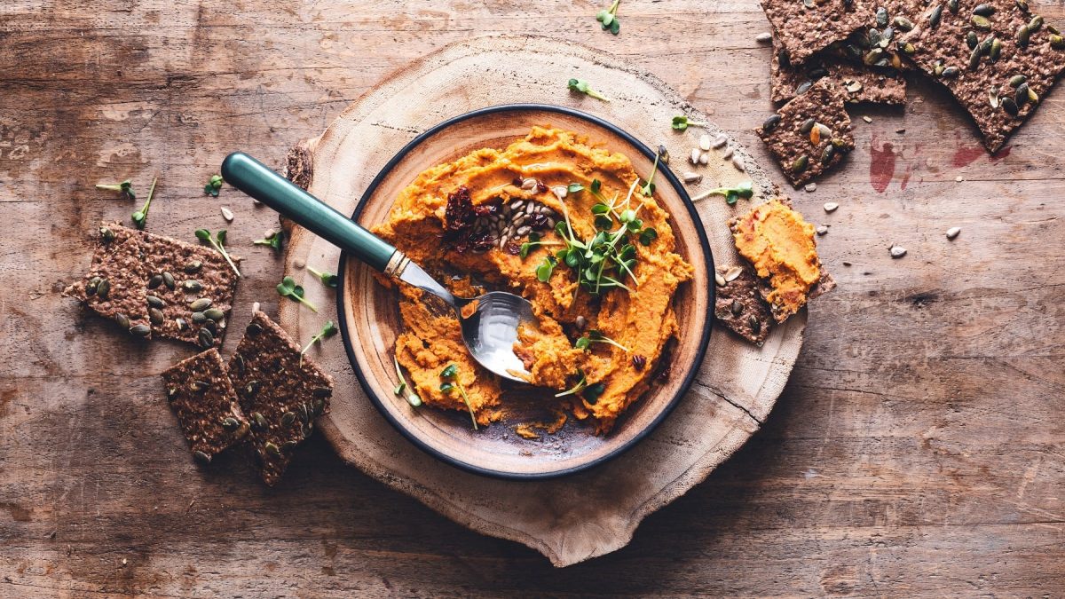 Eine Schüssel Kürbis-Hummus mit Kräutern und Kernen, daneben Cracker, alles Draufsicht.