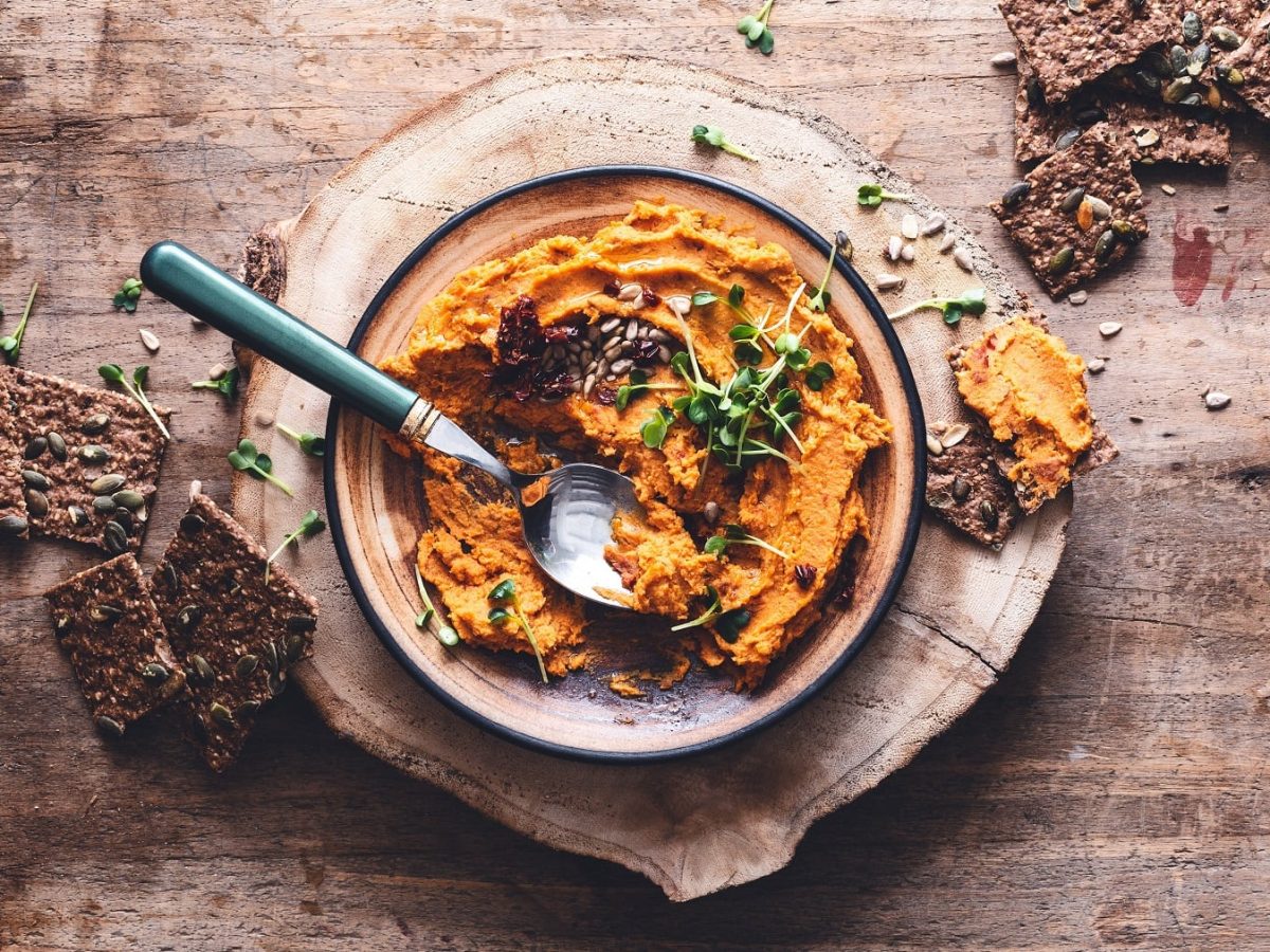 Eine Schüssel Kürbis-Hummus mit Kräutern und Kernen, daneben Cracker, alles Draufsicht.