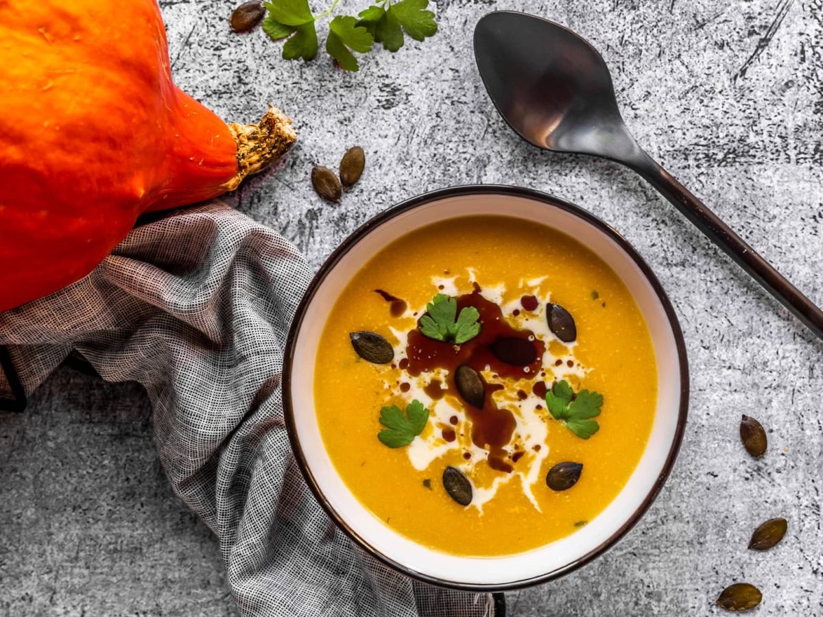 Eine weiße Schale mit Kürbissuppe auf grauem Untergrund. Verfeinert mit Kürbiskernöl, Kürbiskernen, Schmand und Petersilie.