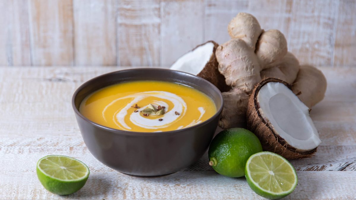 Eine Schale mit Kürbissuppe auf einem weißen Tisch mit Kokos und Limette.