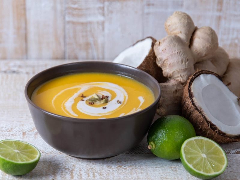 Eine Schale mit Kürbissuppe auf einem weißen Tisch mit Kokos und Limette.