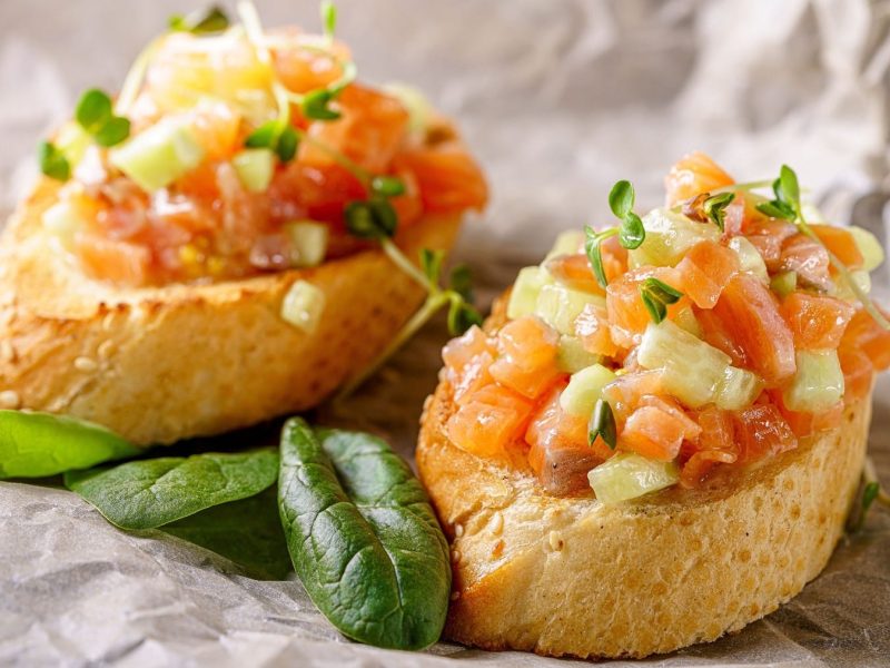 Lachs-Crostinis mit einem Tartar aus Lachswürfeln und Gurkenstückchen auf angerösteten Crostinis, garniert mit frischer Kresse und Basilikum auf hellem Backpapier.