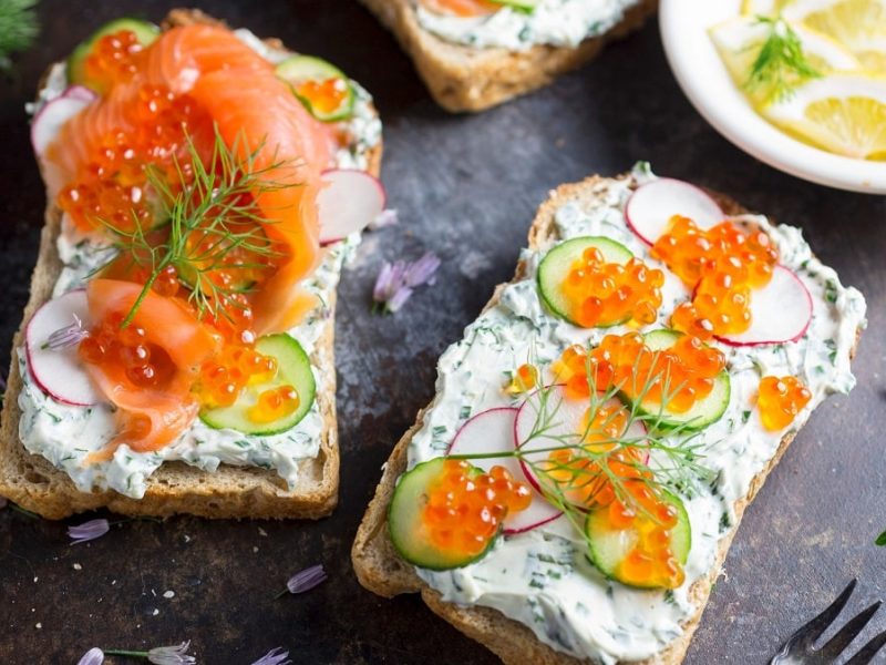 Draufsicht: ein Lachs-Toast mit Forellenkaviar und Radieschen.. Daneben liegt ein Toast mit Forellenkaviar und Radieschen. Drumherum liegen noch frischer Dill, Petersilie und Zitronenscheiben.