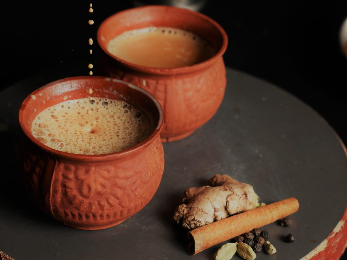 Masala Chai in zwei Tontassen auf dunkler Platte. Daneben Chai Gewürze vor dunklem Hintergrund. Frontalaufnahme.