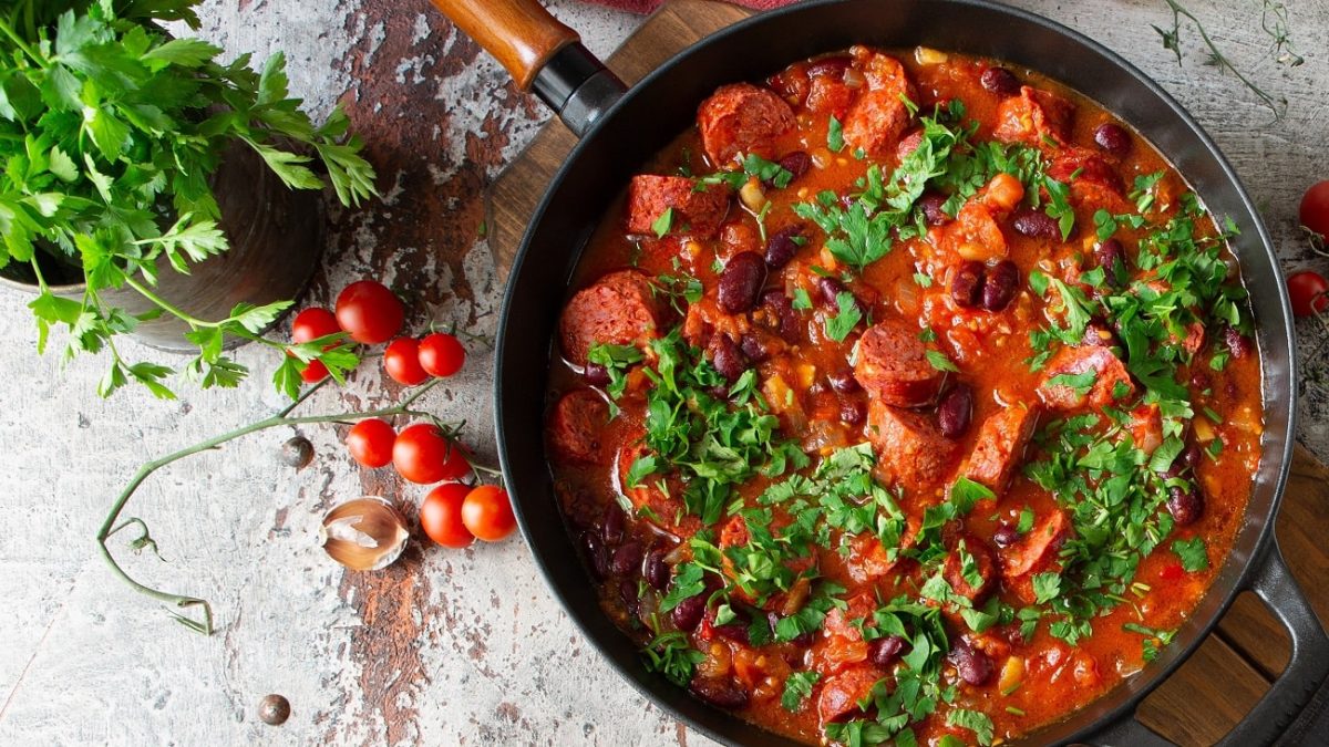 Draufsicht: eine Merguez-Pfanne mit Kidneybohnen und Koriander steht auf einem hellen Untergund. Drumherum liegen kleine Tomatem.