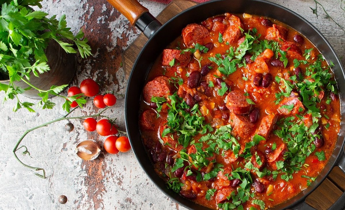 Draufsicht: eine Merguez-Pfanne mit Kidneybohnen und Koriander steht auf einem hellen Untergund. Drumherum liegen kleine Tomatem.