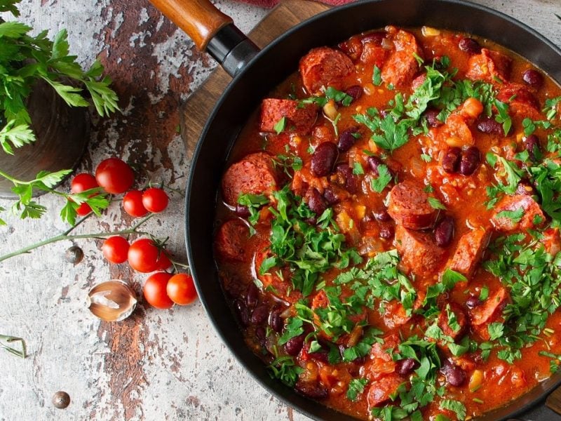 Draufsicht: eine Merguez-Pfanne mit Kidneybohnen und Koriander steht auf einem hellen Untergund. Drumherum liegen kleine Tomatem.