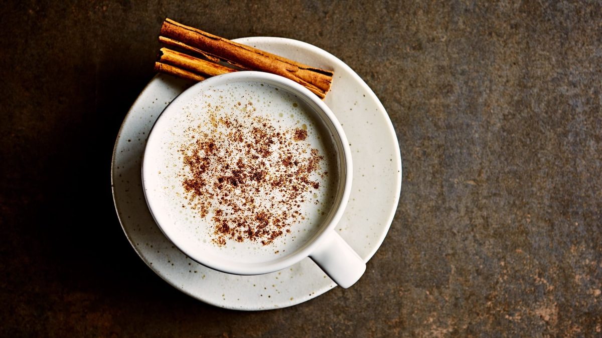 Eine weiße Tasse mit der Mondmilch auf einem dunkelbraunen Untergrund von oben fotografiert. Auf der Untertasse liegen zwei Zimtstangen.