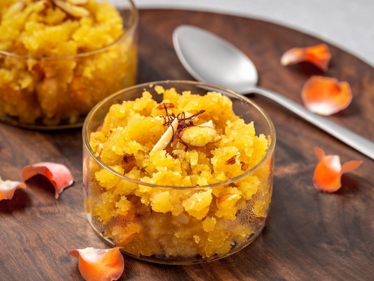 Moong Dal Halwa aus gelben Mungbohnen in Glasschalen auf Holzbrett. Daneben Blüten und Löffel. Draufsicht.