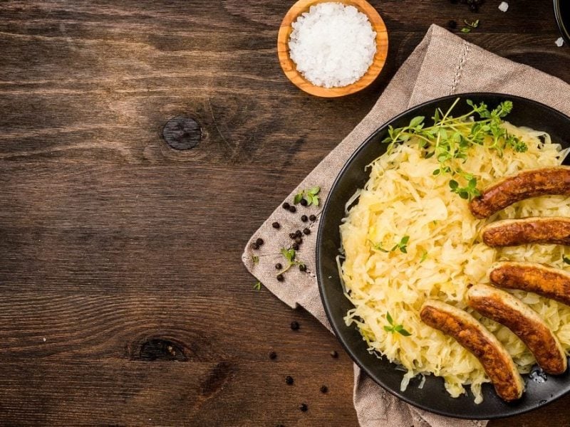 Draufsicht: Ein Teller mit Nürnberger Würstchen mit Sauerkraut steht auf einem Holzbrettchen. Daneben stehen zwei Schalen. Eine mit Salz und eine mit Senf.