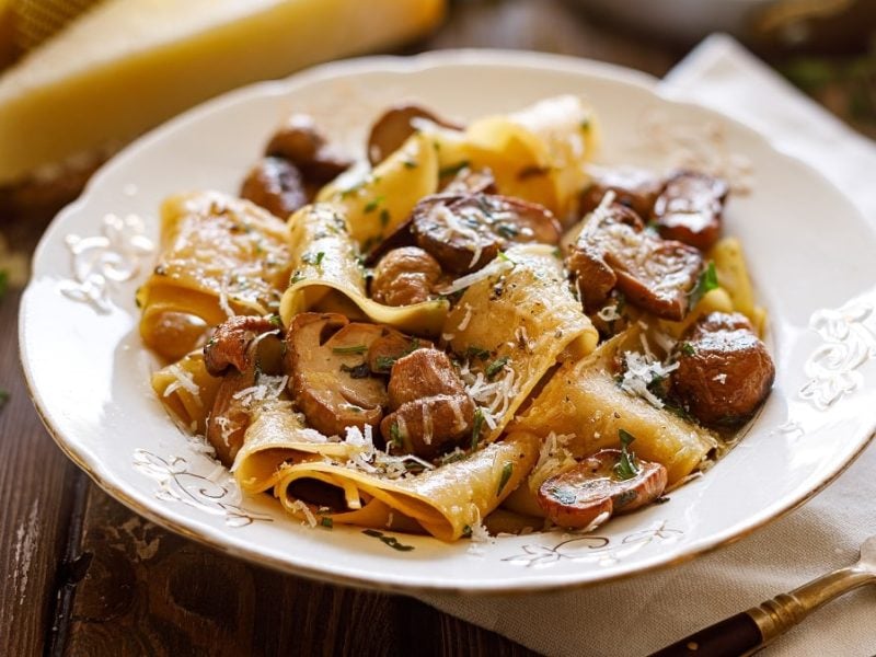 Draufsicht: Ein heller Porzellanteller Pappardelle mit Steinpilzen und Trüffelöl steht auf einem Untergrund auch Holz. Daneben liegen eine Gabel und ein stück Parmesan.