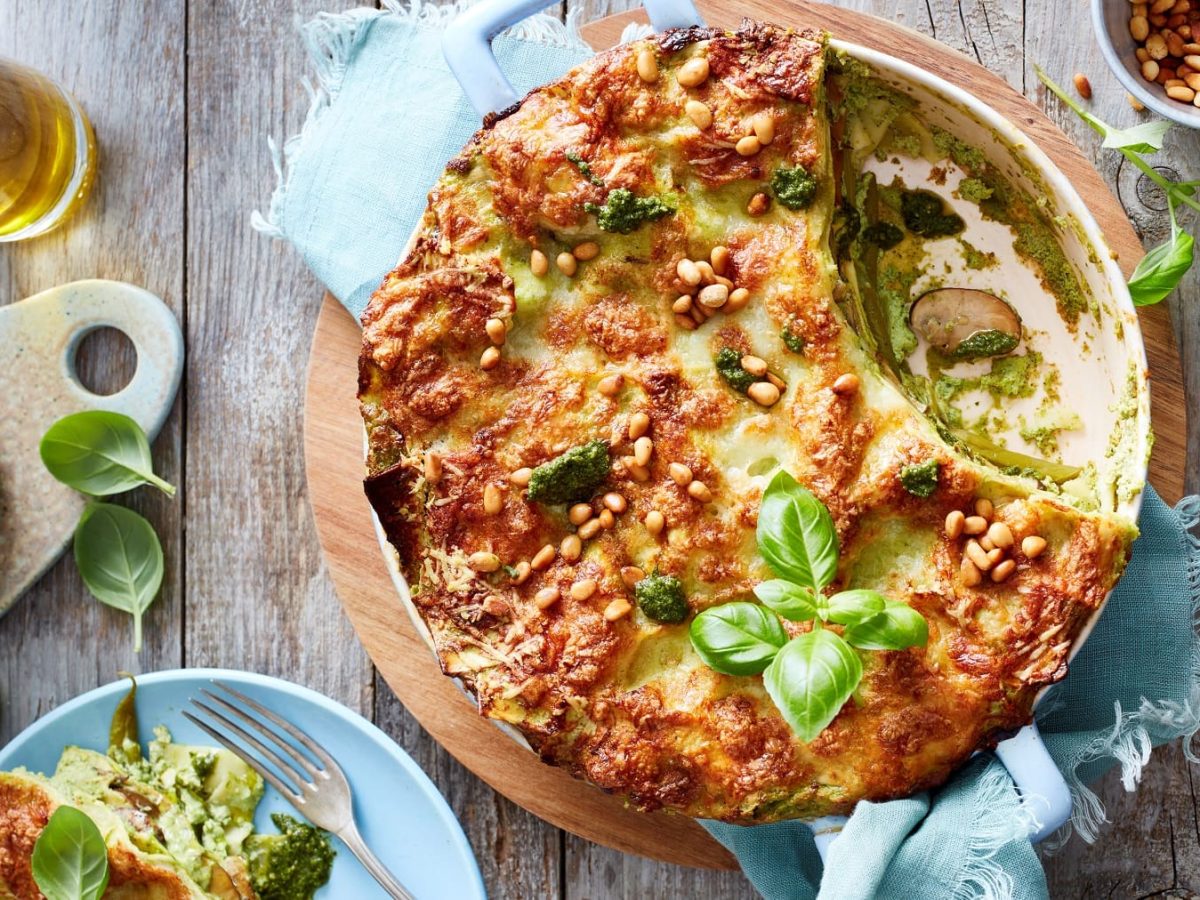 Runde Pesto-Lasagne in der Form neben verschiedenen Zutaten vor Holzhintergrund.