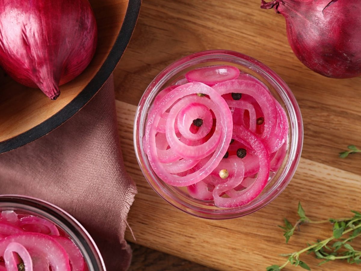 Pickled Red onions in einem Glas neben ganzen roten Zwiebeln und Thymianzweigen vor Holzhintergrund.