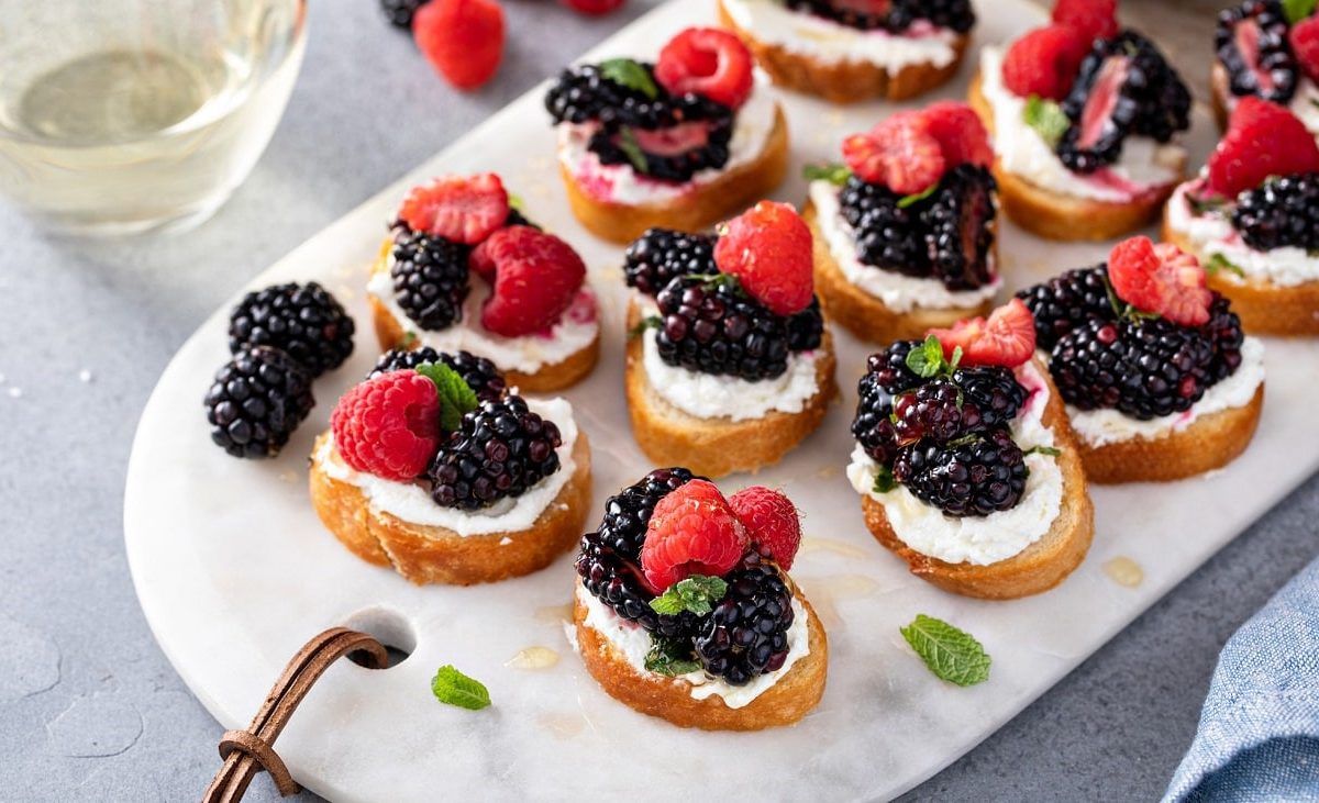 Draufsicht: Auf einem Marmosbrettchen liegen Ricotta-Bruschetta mit Beeren.