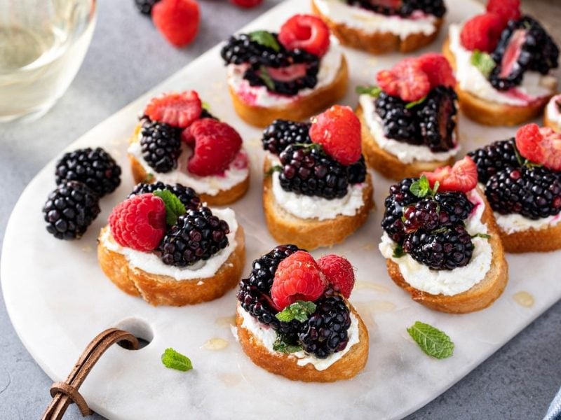 Draufsicht: Auf einem Marmosbrettchen liegen Ricotta-Bruschetta mit Beeren.