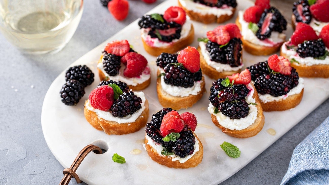 Draufsicht: Auf einem Marmosbrettchen liegen Ricotta-Bruschetta mit Beeren.