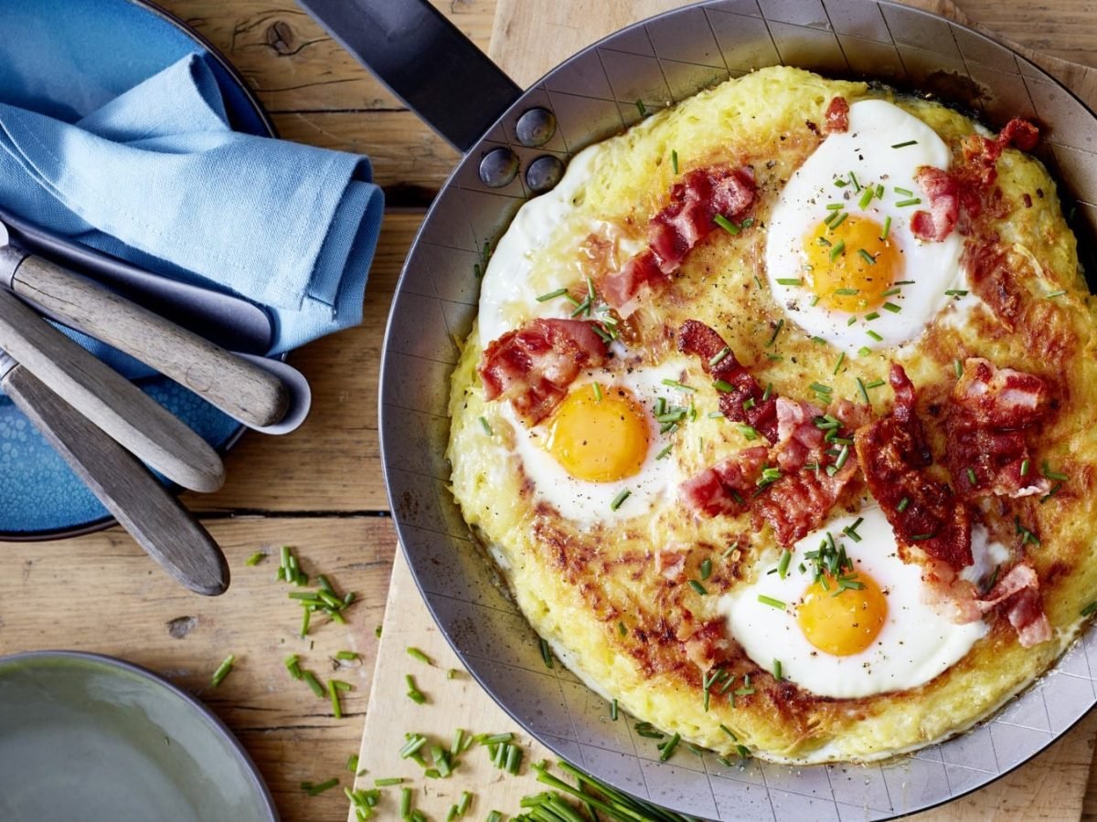 Rösti mit Spiegelei, Bacon und Schnittlauch in einer Pfanne neben Teller und Besteck vor Holzhintergrund.