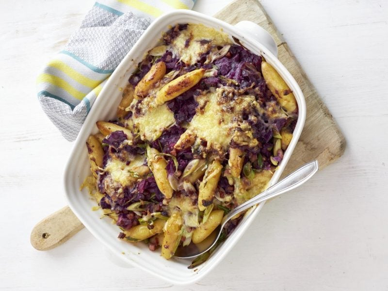 Rotkohl-Schupfnudel-Gratin in weißer Auflaufform auf einem Holzbrett vor hellem Hintergrund.