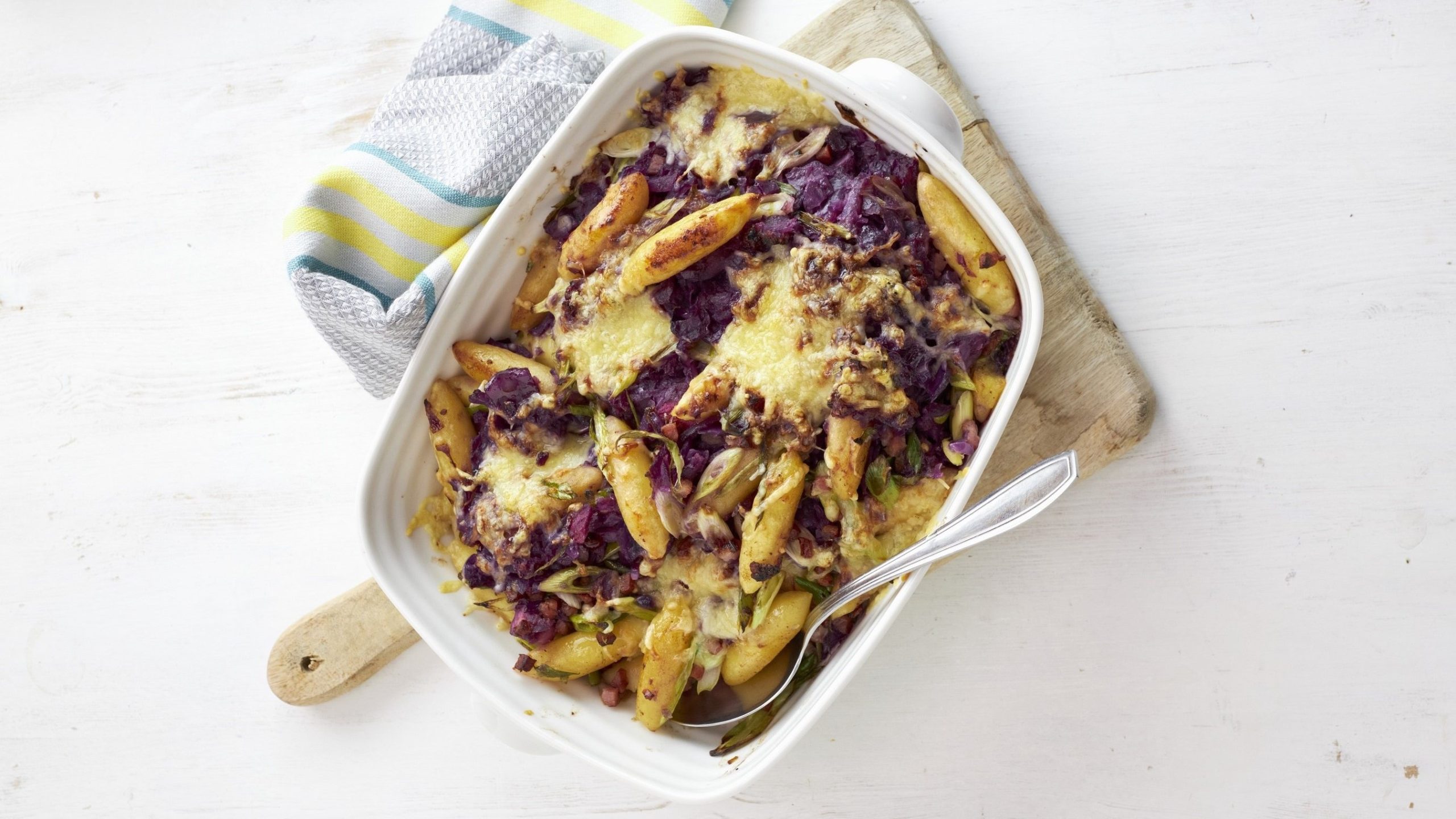 Rotkohl-Schupfnudel-Gratin in weißer Auflaufform auf einem Holzbrett vor hellem Hintergrund.