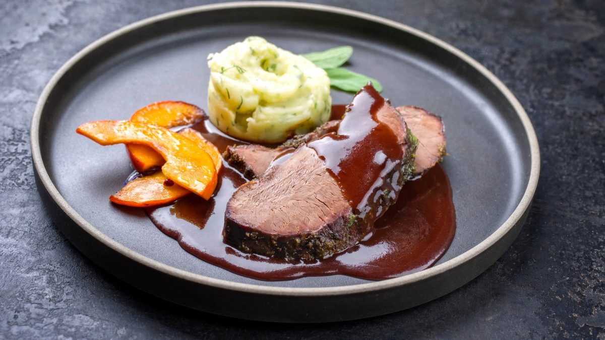 Rosa Hirschfilet mit Kürbis, Kartoffelstampf und Burgundersauce auf grauem Teller mit Salbei auf grauem Untergrund. Draufsicht.