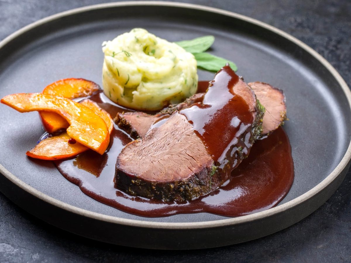 Rosa Hirschfilet mit Kürbis, Kartoffelstampf und Burgundersauce auf grauem Teller mit Salbei auf grauem Untergrund. Draufsicht.