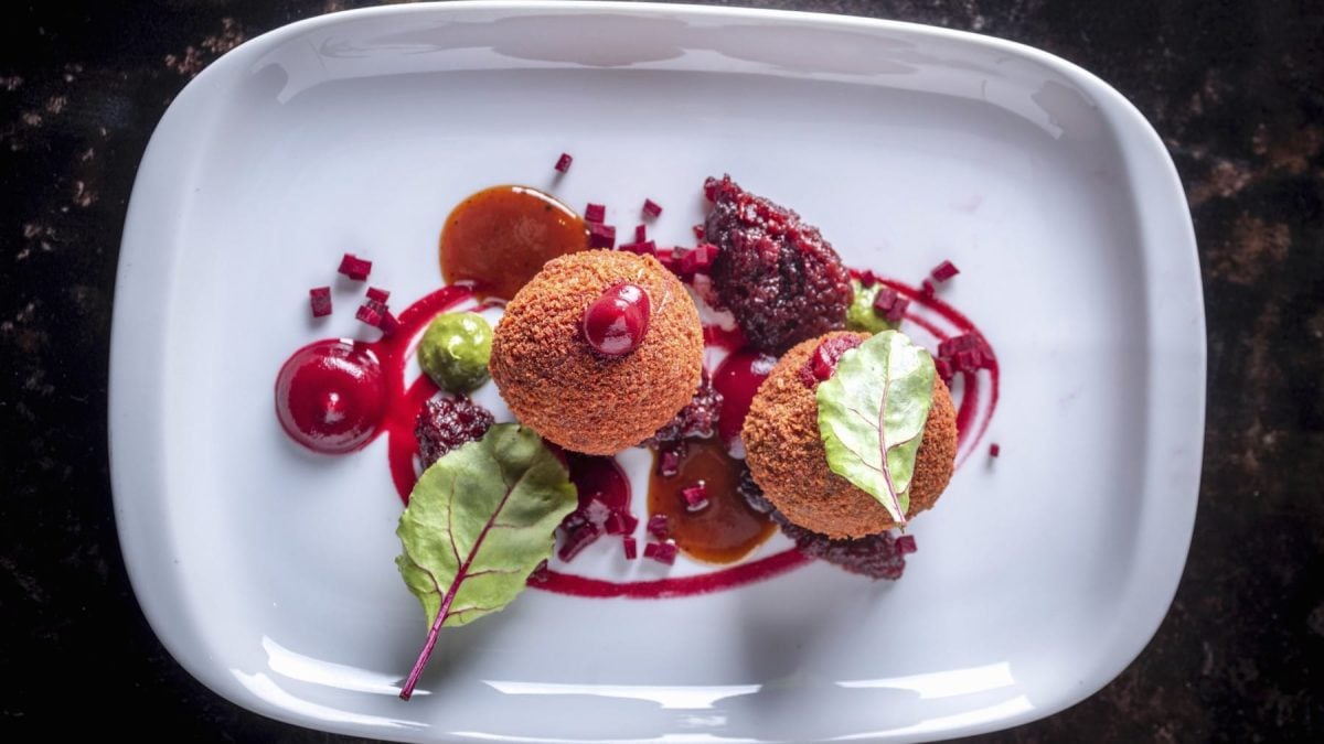 Zwei Rote-Bete-Bällchen auf einem kunstvoll angerichteten weißen, eckigen Teller vor schwarzem Hintergrund.