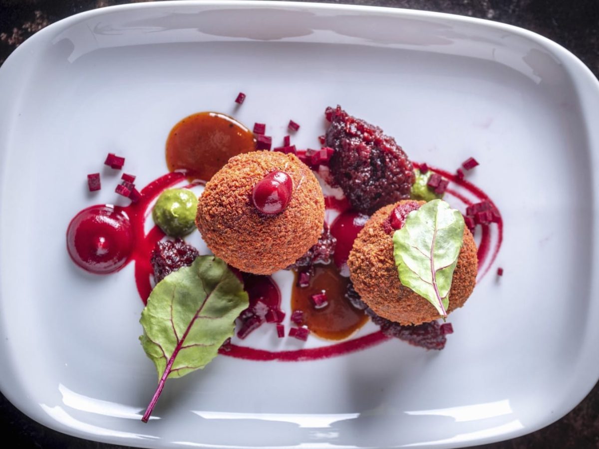 Zwei Rote-Bete-Bällchen auf einem kunstvoll angerichteten weißen, eckigen Teller vor schwarzem Hintergrund.