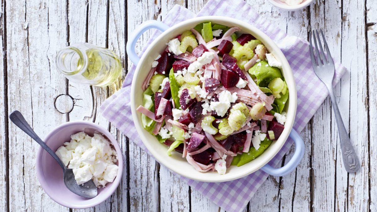 Salat mit Rote Bete und Schinken, garniert mit Schafskäse in einer weißen Schüssel mit hellem Holzhintergrund.