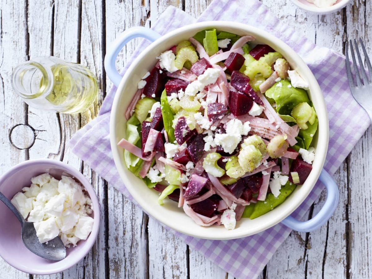 Salat mit Rote Bete und Schinken, garniert mit Schafskäse in einer weißen Schüssel mit hellem Holzhintergrund.