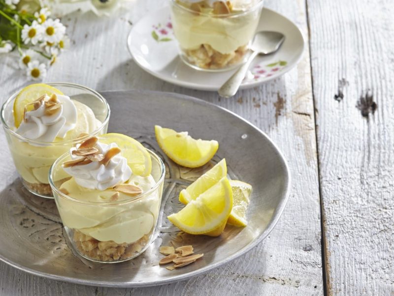 Schichtdessert mit Lemoncurd und Limoncello in Gläsern auf weißem Teller mit Zitronenspalten. Daneben Blumendekoration. Draufsicht.