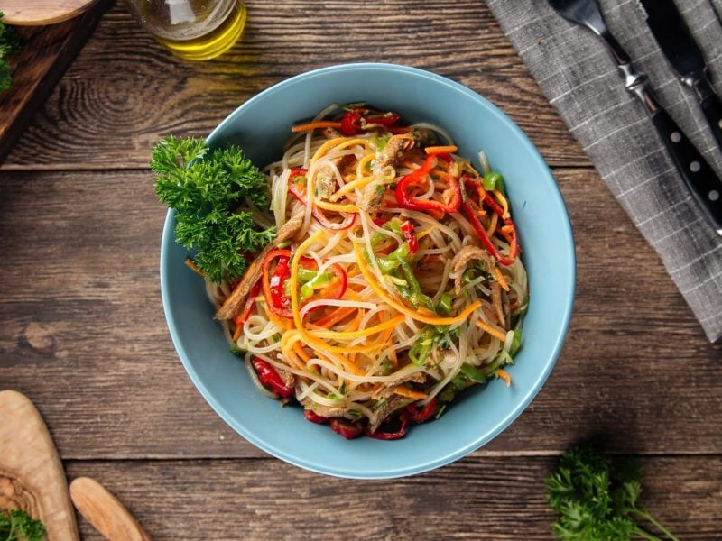 eine Schüssel schnelle asiatische Pasta mit Erdnusssauce und bunter Paprika sowie Petersilie in der Draufsicht, daneben Besteck.