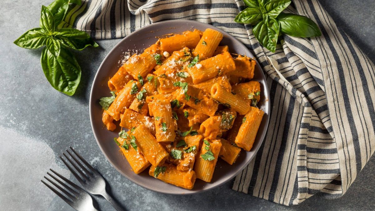 Ein Teller schnelle Paprika-Pasta, Penne, mit Basilikum und Parmesan bestreut, in der Draufsicht. Daneben ein Geschirrtuch, zwei Gabeln und Basilikum.