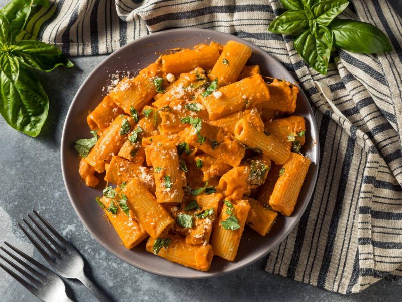 Ein Teller schnelle Paprika-Pasta, Penne, mit Basilikum und Parmesan bestreut, in der Draufsicht. Daneben ein Geschirrtuch, zwei Gabeln und Basilikum.