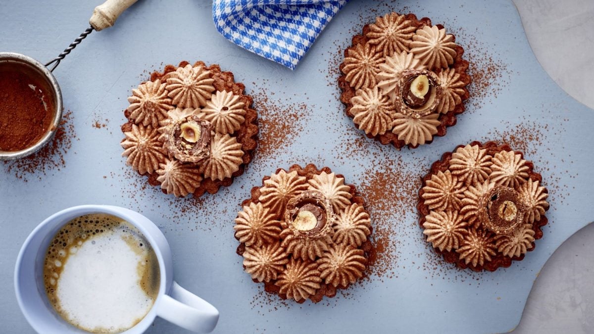 Vier Schoko-Tartelettes mit Frischkäsecreme neben einer Tasse Kaffee und Kakaopulver im Sieb vor blauem Hintergrund.