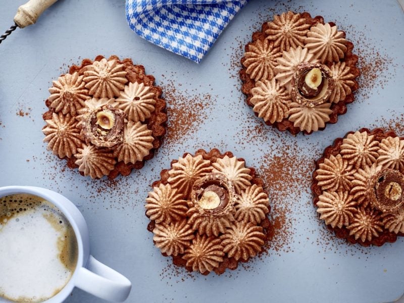 Vier Schoko-Tartelettes mit Frischkäsecreme neben einer Tasse Kaffee und Kakaopulver im Sieb vor blauem Hintergrund.