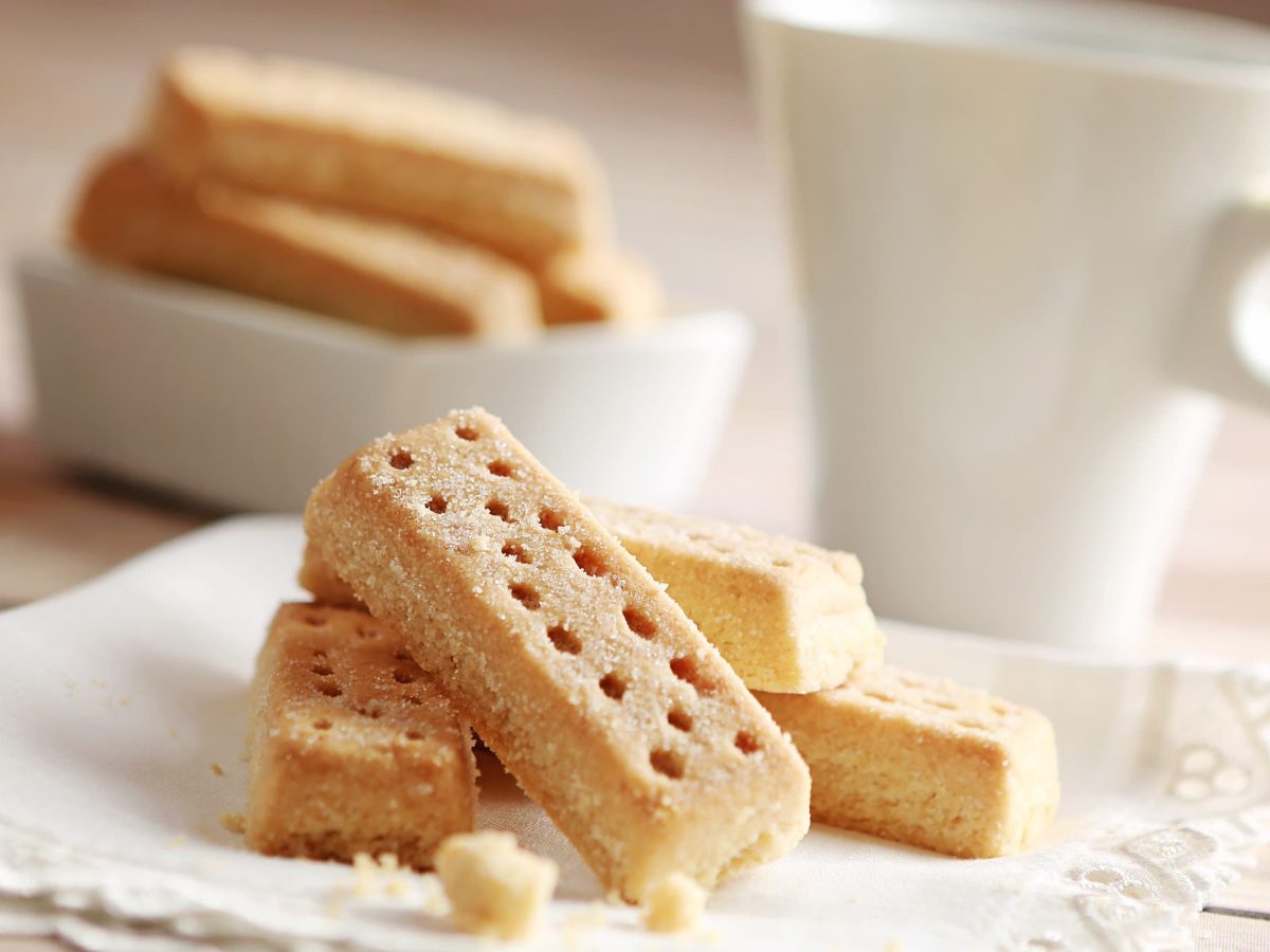 Schottisches Shortbread auf Stoffservierte und hellem Untergrund. Im Hintergrund eine weiße Tasse und eine Schale mit weiteren Shortbread. Frontalansicht.