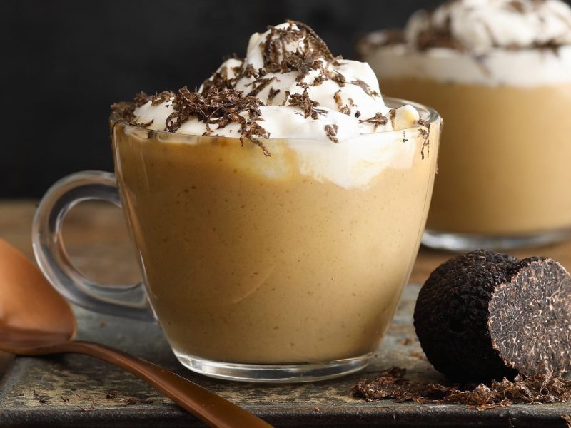 Zwei Glastassen Steinpilz-Cappuccino mit Trüffelsahne, daneben Löffel und Trüffel vor dunklem Hintergrund.