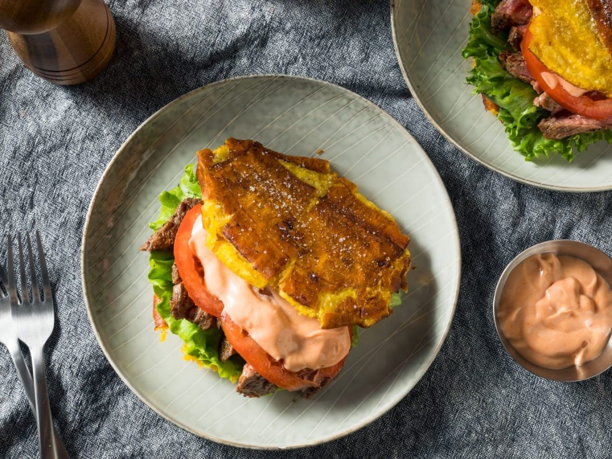 Zwei Tostones Burger auf zwei hellen Tellern neben einer Schale mit Sauce, einer Gabel und einer Gewürzmühle vor grauem Stoffhintergrund.