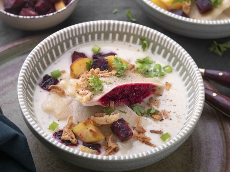 Vegane Maultaschen mit Rote-Bete-Füllung in heller Sauce in einer Schüssel vor dunklem Hintergrund.