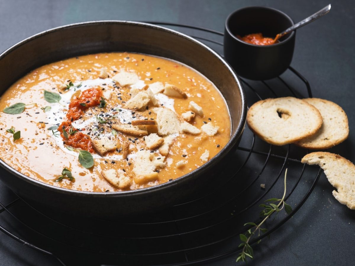 Vegane Paprika-Kartoffel-Suppe in einer schwarzen Schüssel neben Knoblauch-Brotchips und Ajvar vor schwarzem Hintergrund.