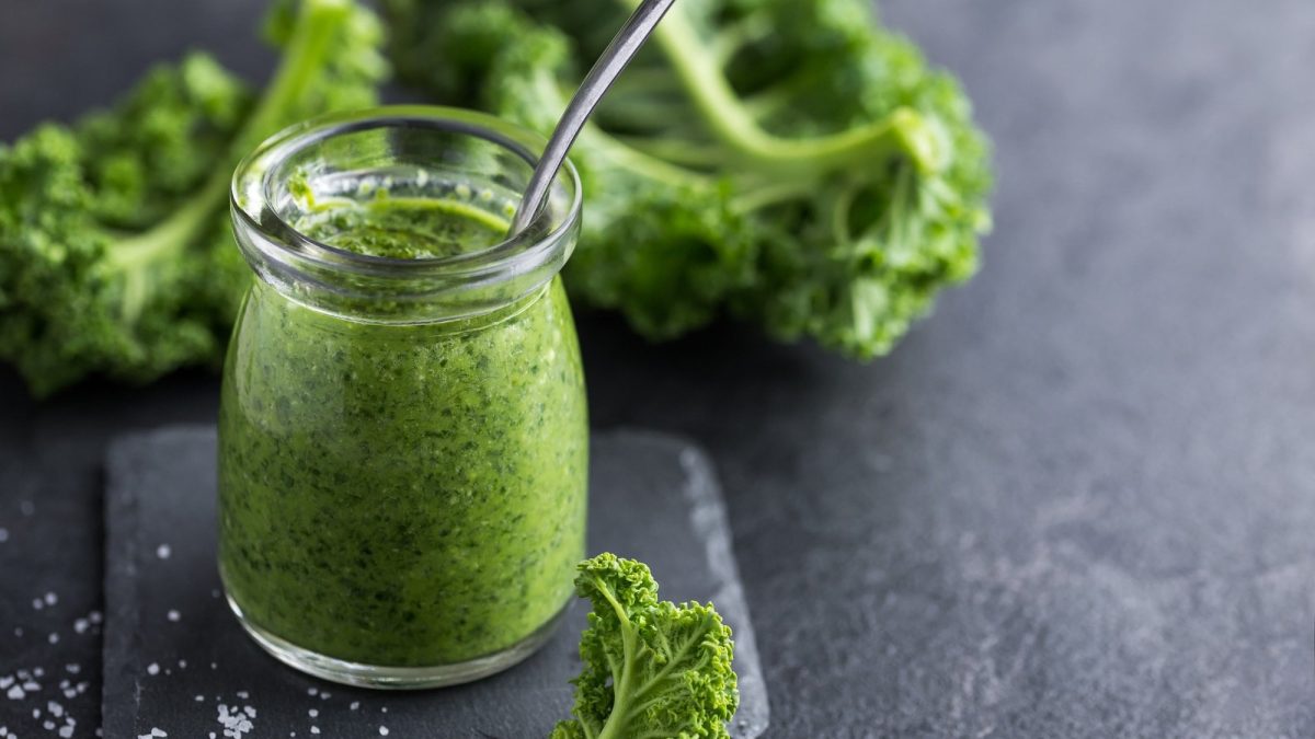 Ein Glas veganes Grünkohlpesto mit einem Löffel, drumherum liegt frischer Grünkohl.