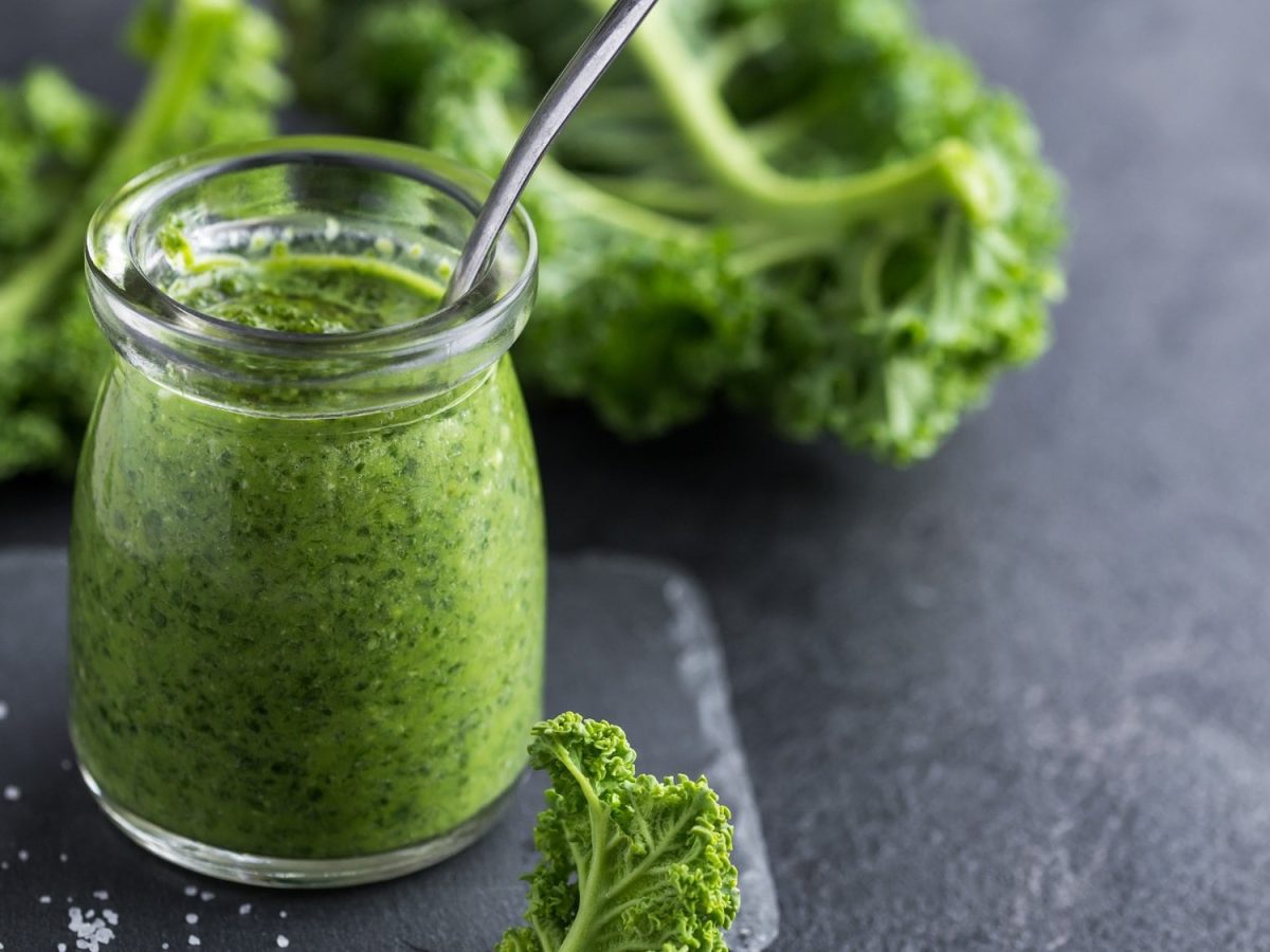 Ein Glas veganes Grünkohlpesto mit einem Löffel, drumherum liegt frischer Grünkohl.