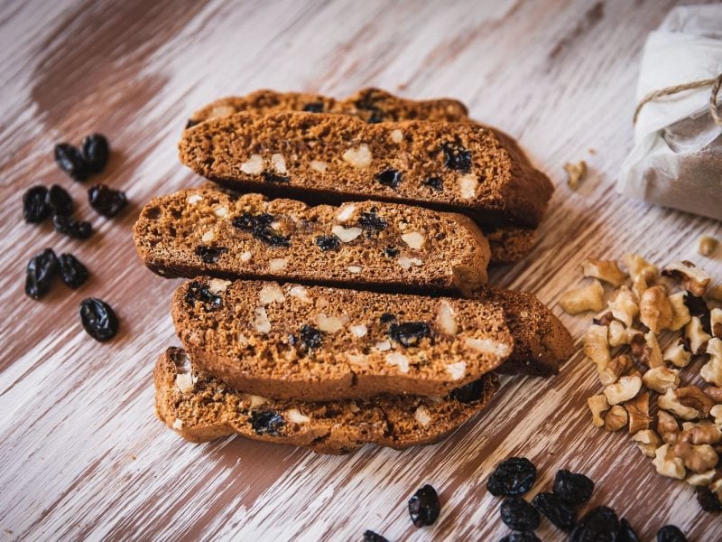 Walnuss-Biscotti auf einem Haufen, daneben Rosinen und gehackte Walnüsse.