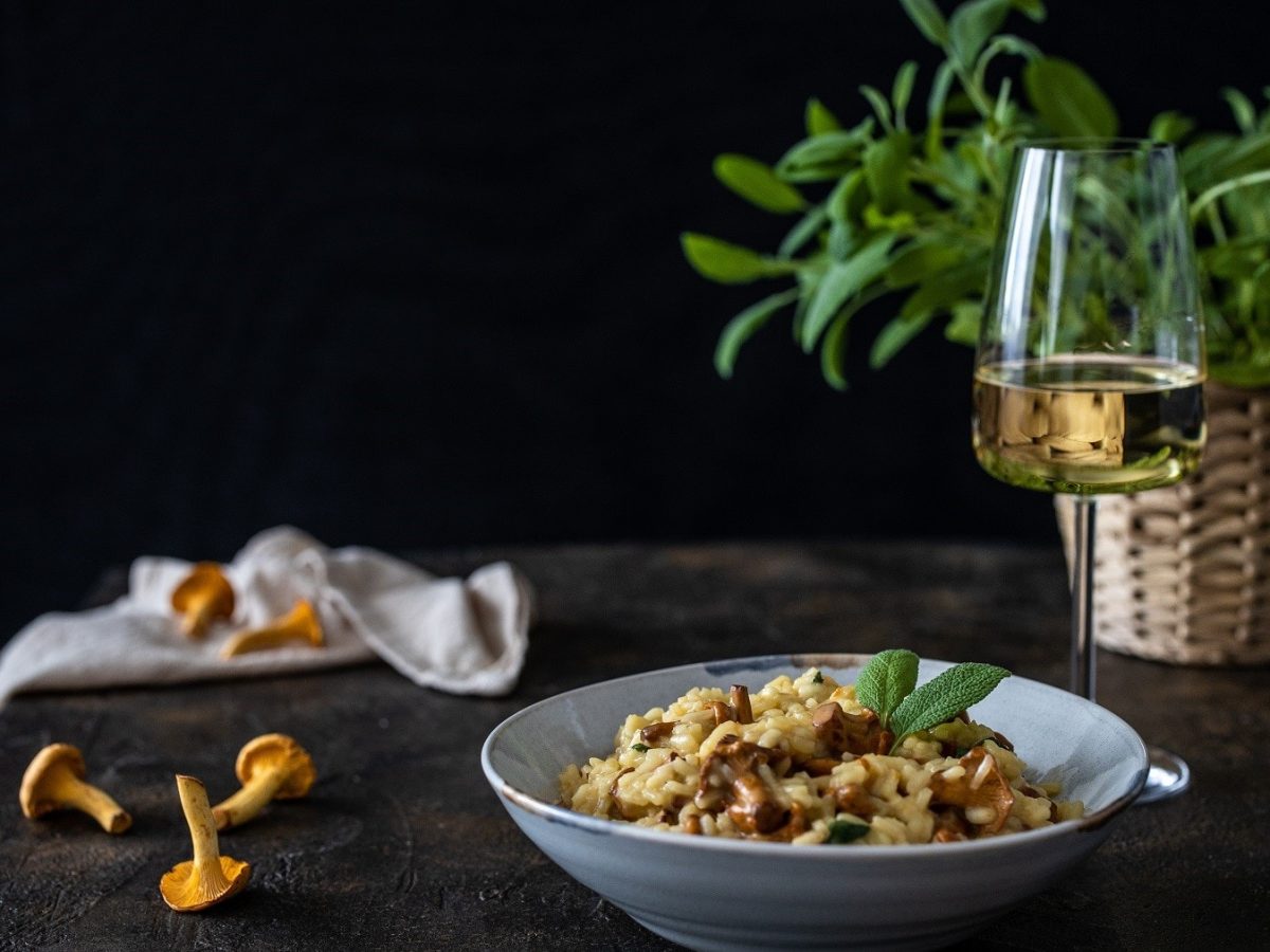 Welcher Wein zu welchem Pilz – so zum Beispiel wie ein milder Weißwein zu dem auf dem Bild servierten Pfifferling-Risotto, auf einem dunklen Tisch mit einem Korb mit frischen Kräutern.