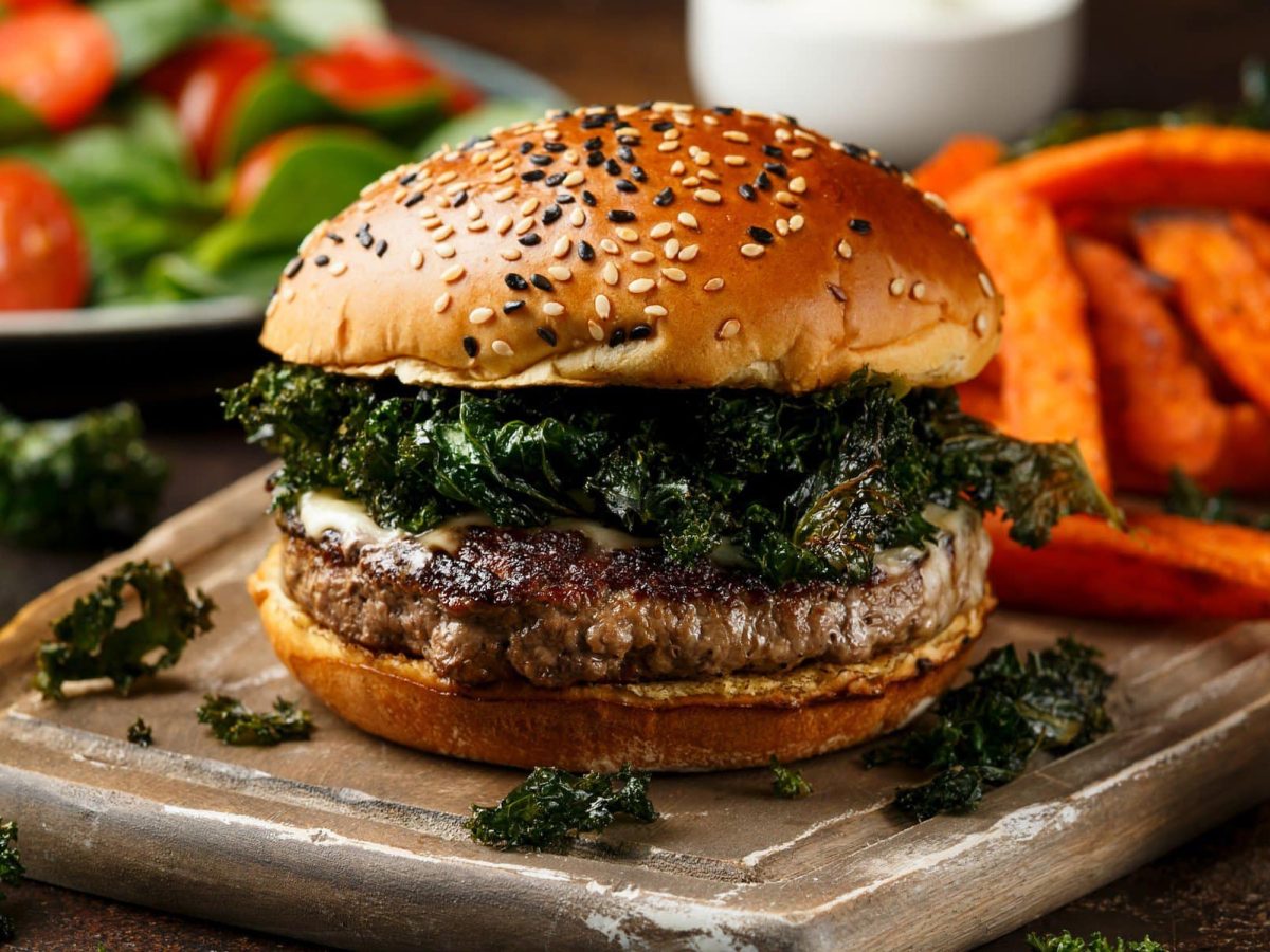 Wildschwein-Burger mit frittiertem Grünkohl auf Anrichtebrett. Im Hintergrund Süßkartoffelpommes, Dips, Salat und ein Glas Bier. Frontalansicht.