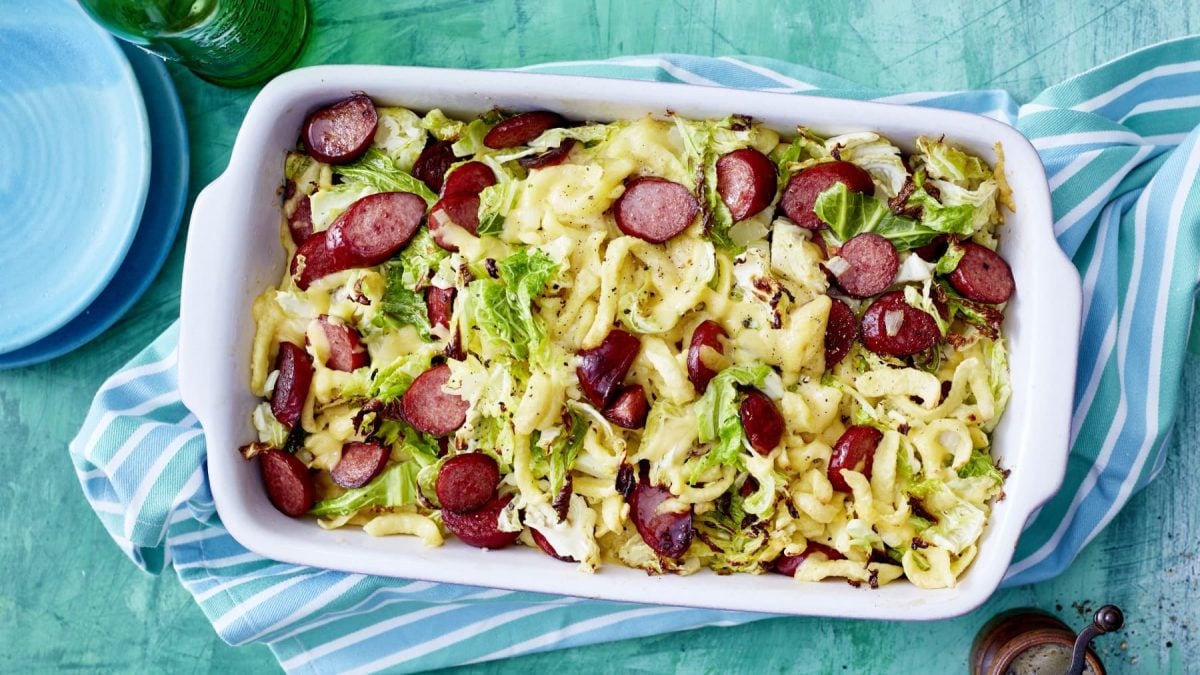 Auflaufform mit Ofen-Käse-Spätzle und Cabanossi vor bläulichem Hintergrund.