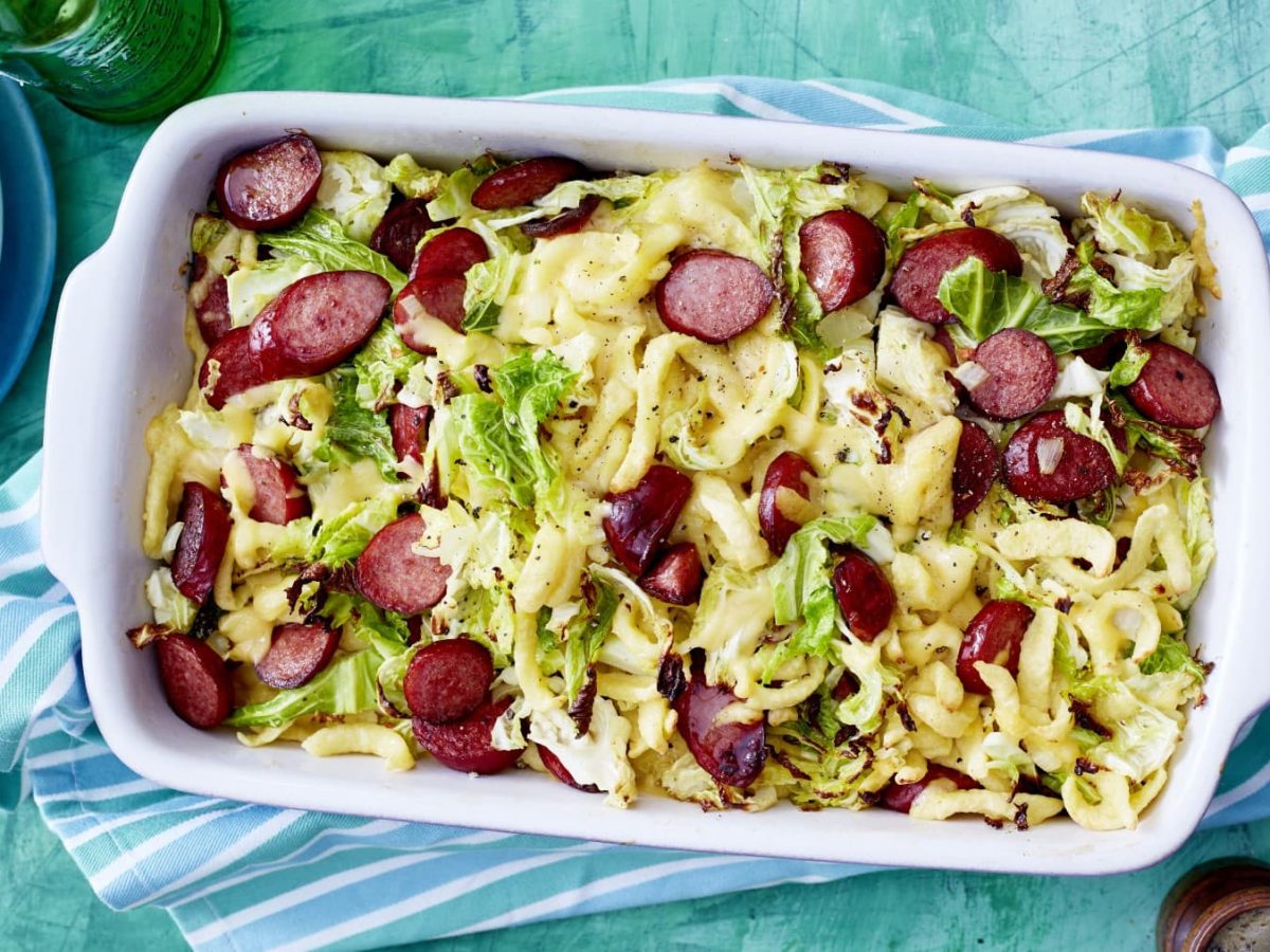 Auflaufform mit Ofen-Käse-Spätzle und Cabanossi vor bläulichem Hintergrund.