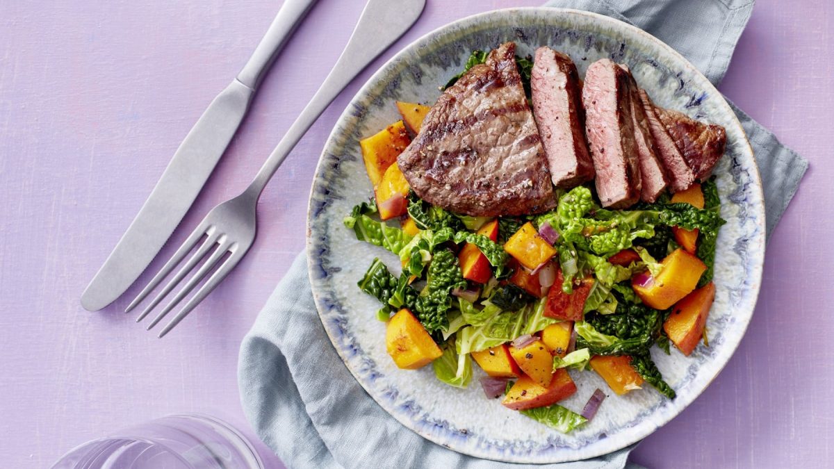 Teller mit Wirsing und Kürbis mit Steak neben Besteck vor lila Hintergrund.