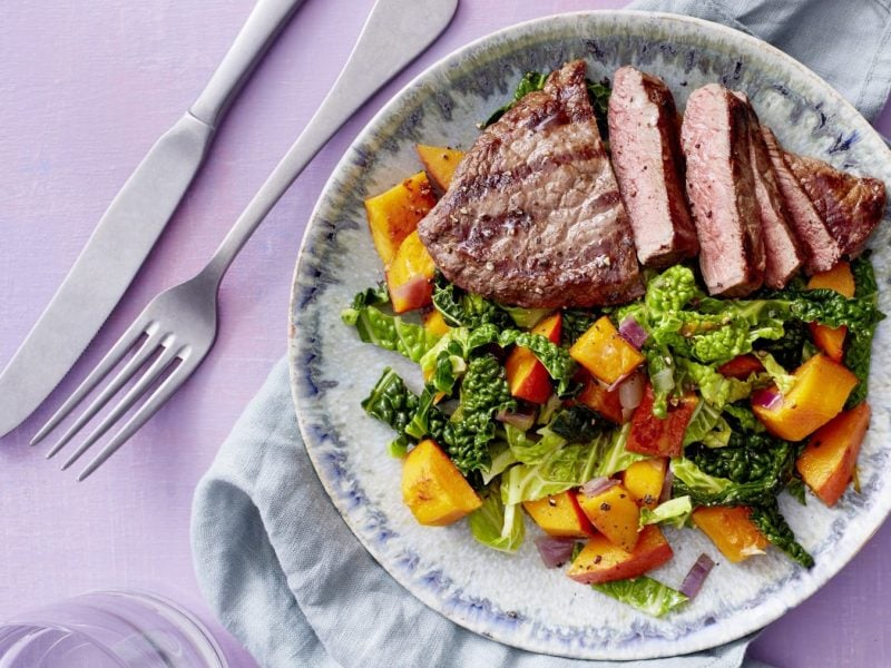 Teller mit Wirsing und Kürbis mit Steak neben Besteck vor lila Hintergrund.